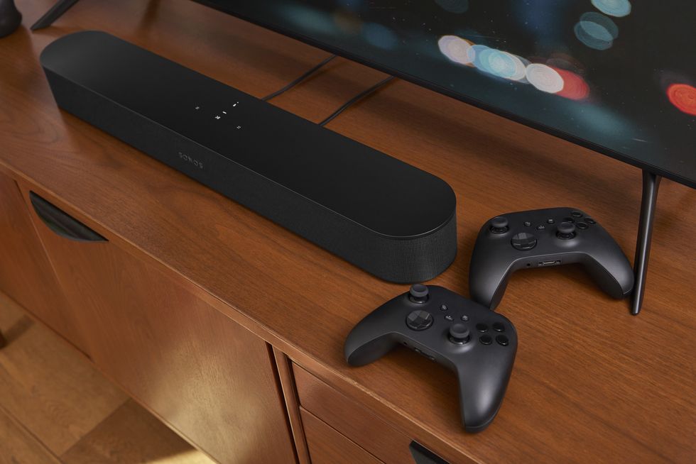 sonos beam gen 2 is pictured on a table with two xbox controllers