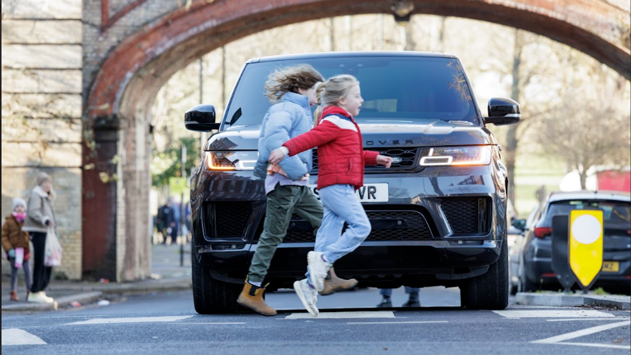 Two children cross a road in front of a large Land Rover SUV