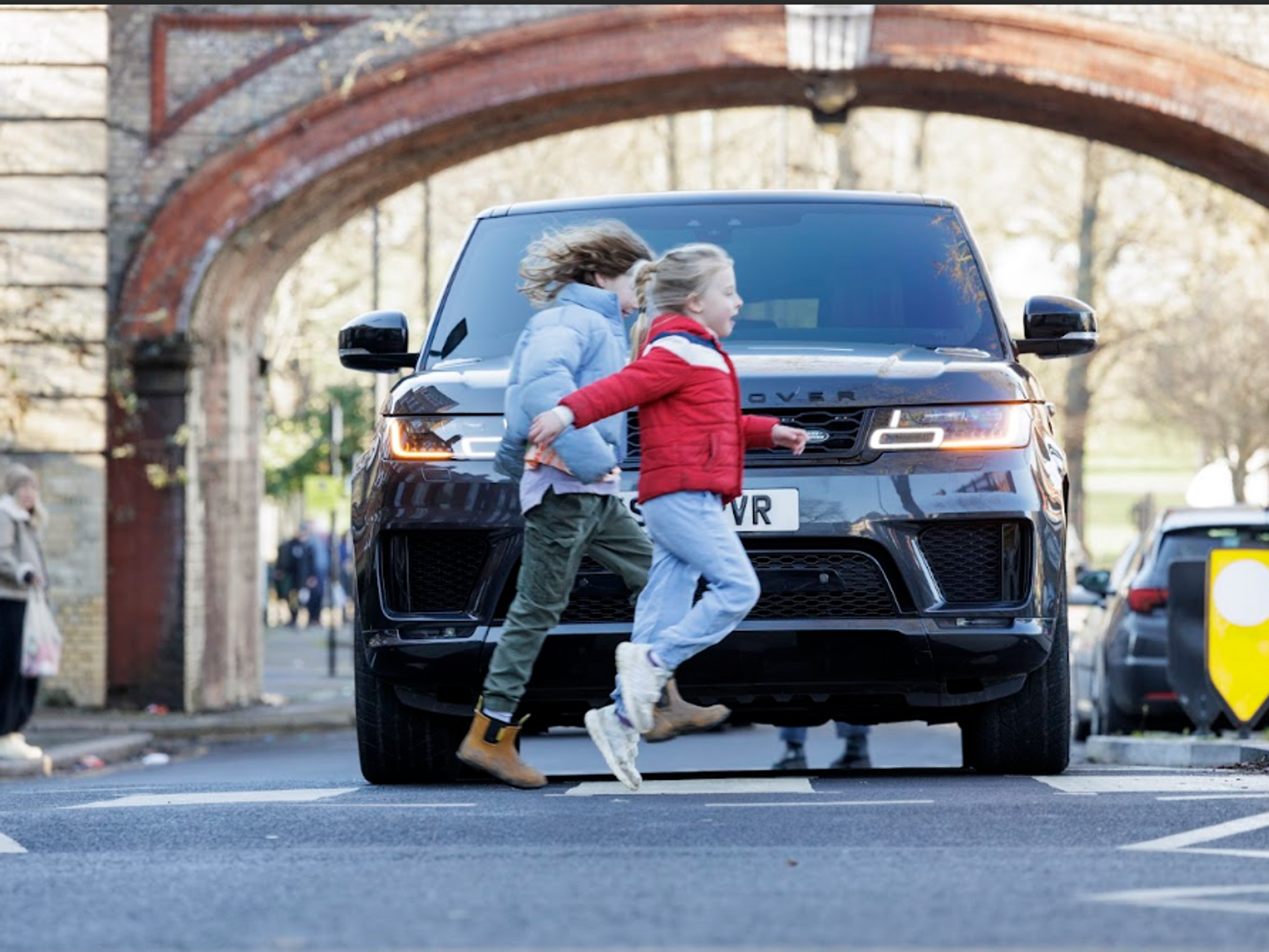 Two children cross a road in front of a large Land Rover SUV