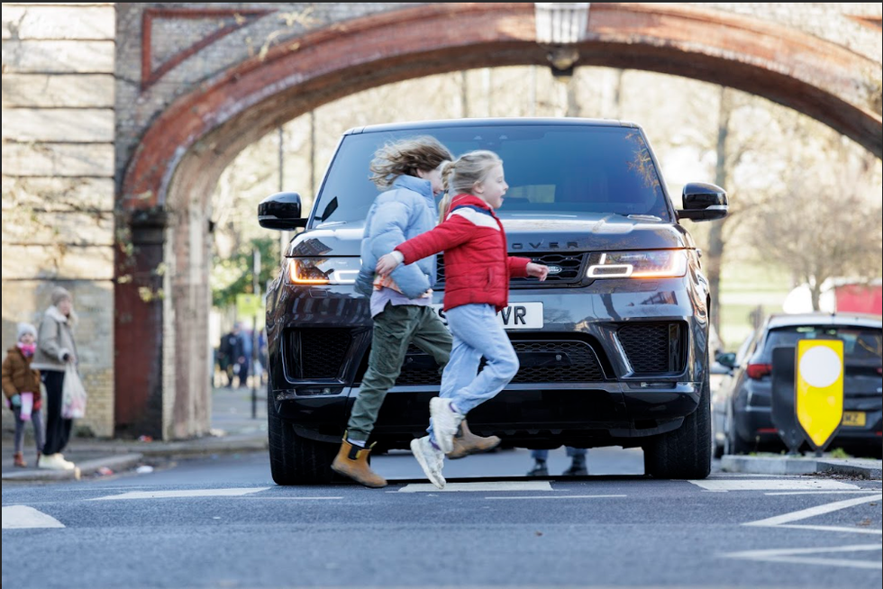 Two children cross a road in front of a large Land Rover SUV