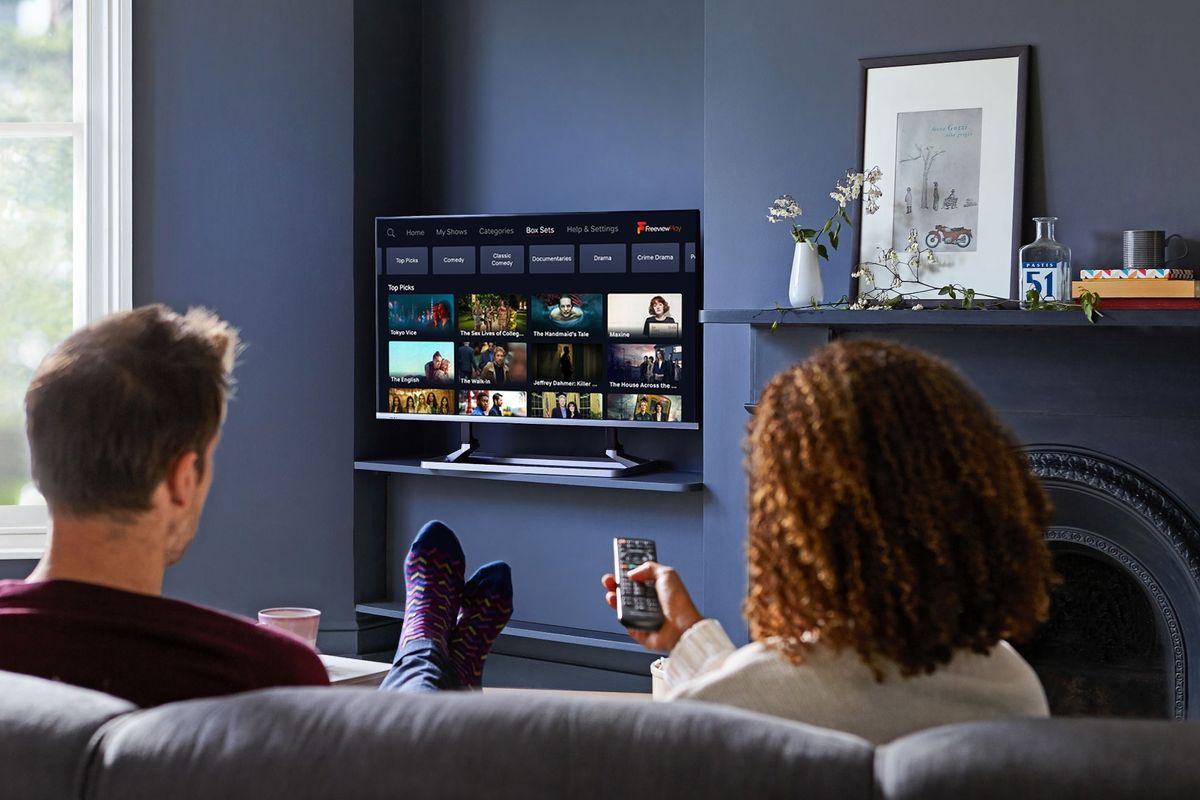 two people sit on a sofa watching a flatscreen television with the freeview menu on-screen 