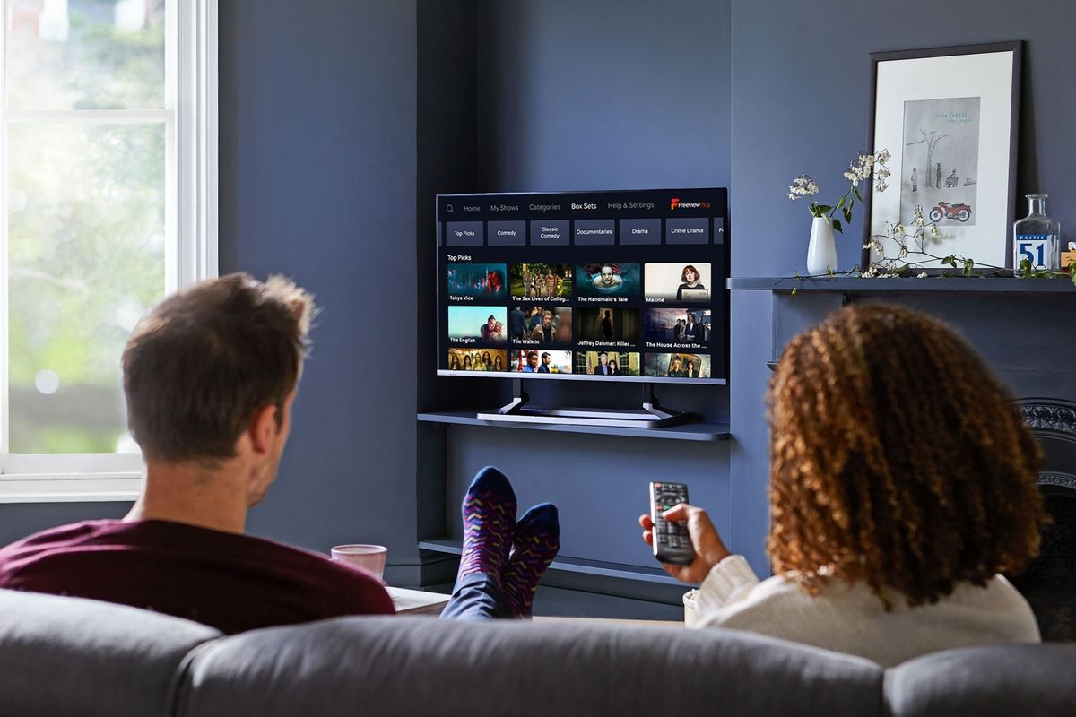 two people sit on a sofa watching a flatscreen television with the freeview menu on-screen