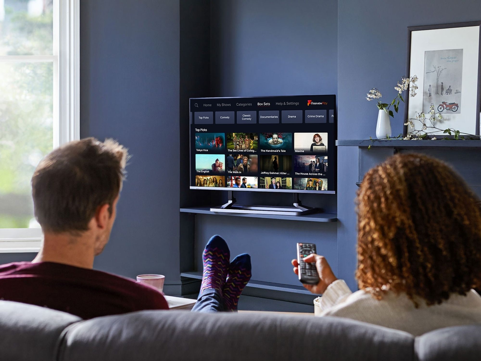 two people sit on a sofa watching a flatscreen television with the freeview menu on-screen
