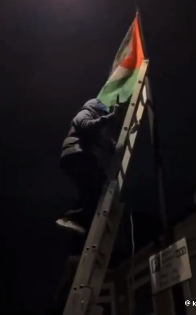 \u200bA protester climbs a ladder to attach a Palestinian flag to a lamppost outside of the school