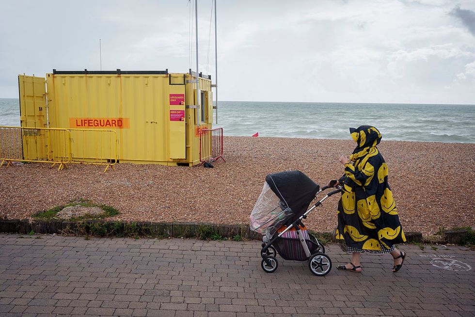 \u200bA woman and a child walk along the seafront, passing a lifeguard hut on the shingle beach at Hove