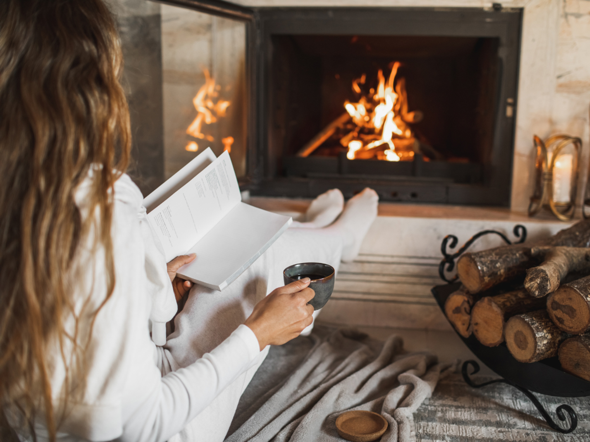 Woman holding a book and cup of tea in front of a fire