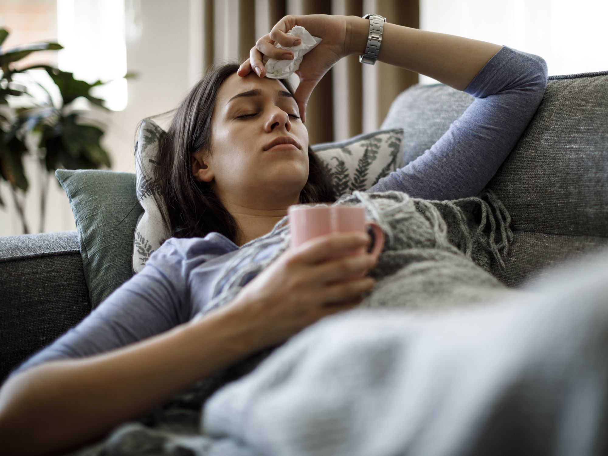 Woman with the flu holding her head and wrapped in a blanket on the sofa