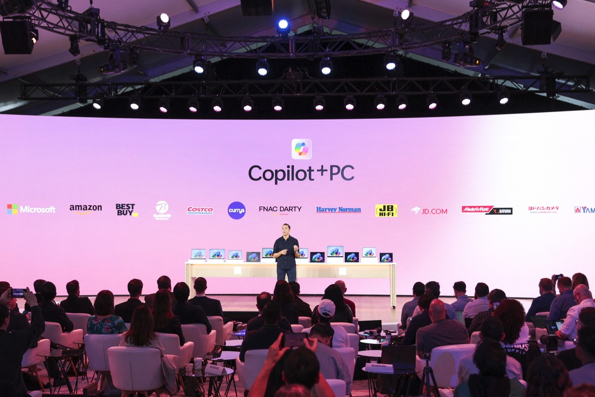 Yusuf Mehdi on-stage with a shelf of laptops running windows 11